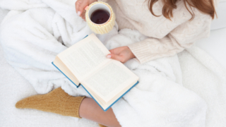 白いブランケットに包まれて本を読みながらホットドリンクを楽しむ女性