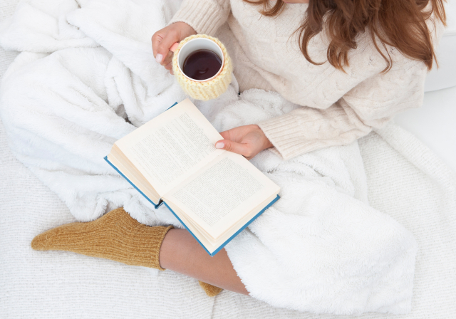 白いブランケットに包まれて本を読みながらホットドリンクを楽しむ女性