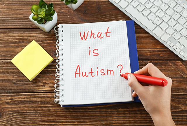 「What is Autism?」とノートに赤ペンで書かれた文字。木目調の机の上にメモ帳や植物、キーボードが置かれている構図。
