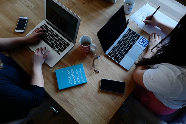 木製のデスクでノートパソコンとメモ帳、スマホを使って共同作業を行う2人の女性。