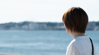 海辺の柵にもたれて海を見つめる白いシャツの女性の後ろ姿