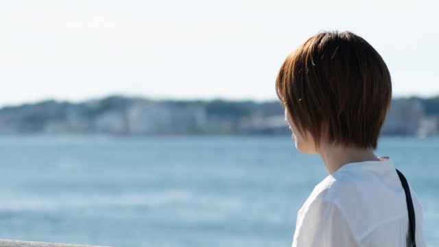 海辺の柵にもたれて海を見つめる白いシャツの女性の後ろ姿