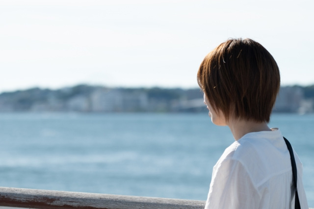 海辺の柵にもたれて海を見つめる白いシャツの女性の後ろ姿
