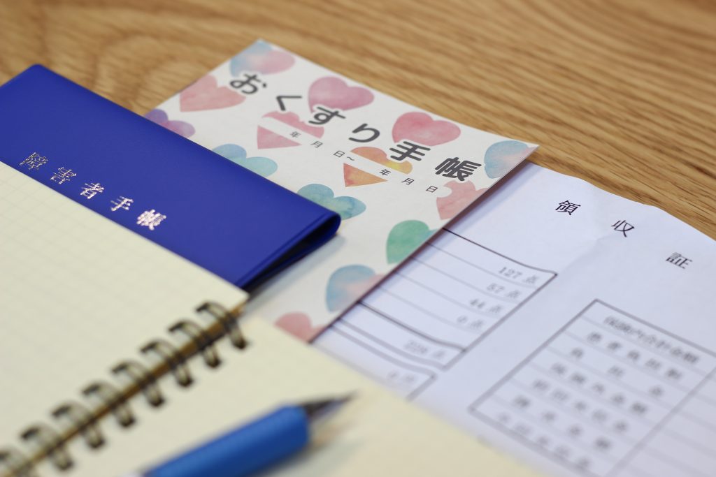 親なき後の生活設計を考えるための「おくすり手帳」や収入証明書、障害年金手帳とノート・ペンのイメージ写真