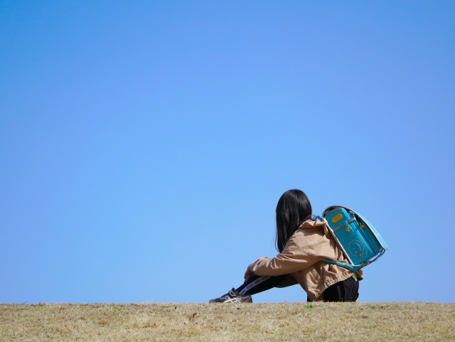 青空の下でリュックを背負い、一人で座って考え込む人物。視覚障害者が将来の職業や生き方について悩み、進路を模索する心情を表している