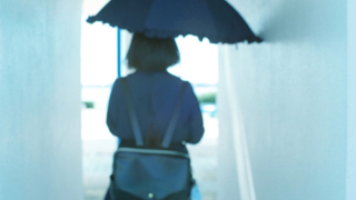 高校2年生でSLEを発症し、診断確定まで不安な日々を過ごす女子高校生の後ろ姿。傘を差しながら狭い通路を歩く姿が病気と向き合う孤独や葛藤を象徴している。