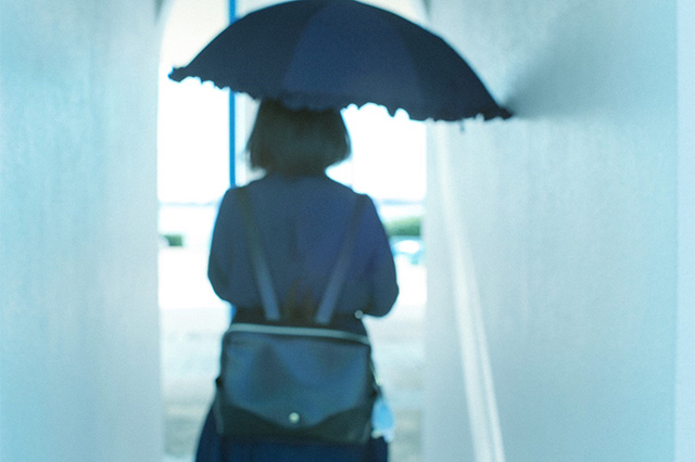 高校2年生でSLEを発症し、診断確定まで不安な日々を過ごす女子高校生の後ろ姿。傘を差しながら狭い通路を歩く姿が病気と向き合う孤独や葛藤を象徴している。