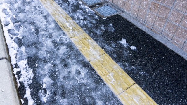 雪と氷で滑りやすくなった歩道の点字ブロック。冬の悪天候が全盲者の外出時に与える危険を示している様子。